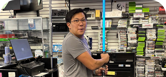 Staff member at Disc Replay posing with a blue lightsaber inside the store’s gaming section